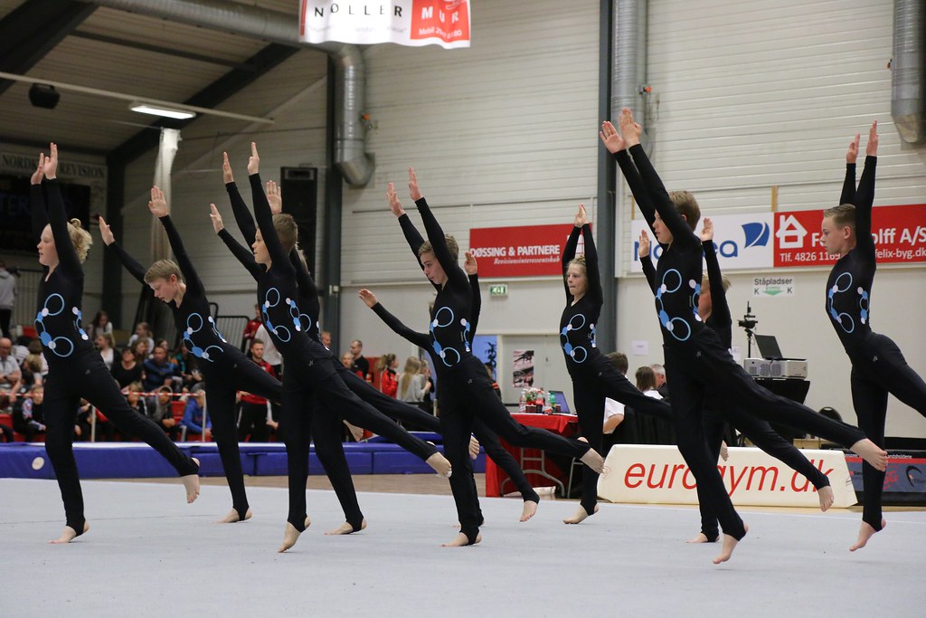 Hold af gymnaster i ens sorte dragter med blå mønstre, der optræder i en sportshal.