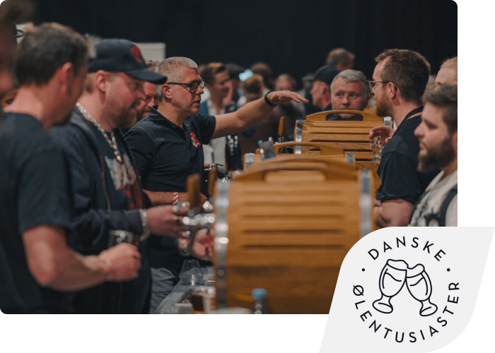 A group of men gathered around a bar with small wooden barrels at a beer tasting. The Danske Ølentusiaster logo is inset in the corner.