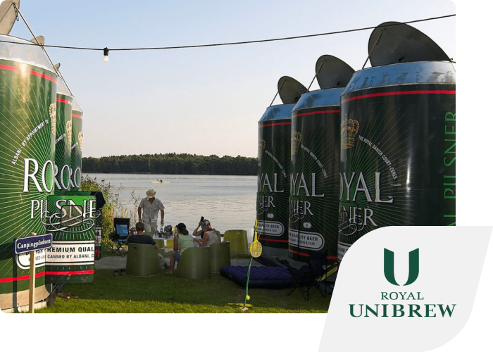 Large beer cans placed on a meadow with seating in between. The Royal Unibrew logo is inset in the corner.