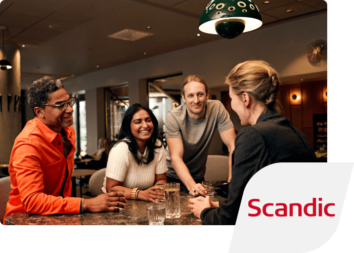 Four smiling people talking over a couple of glasses of water in a cosy lounge. The Scandic logo is inset in the corner.